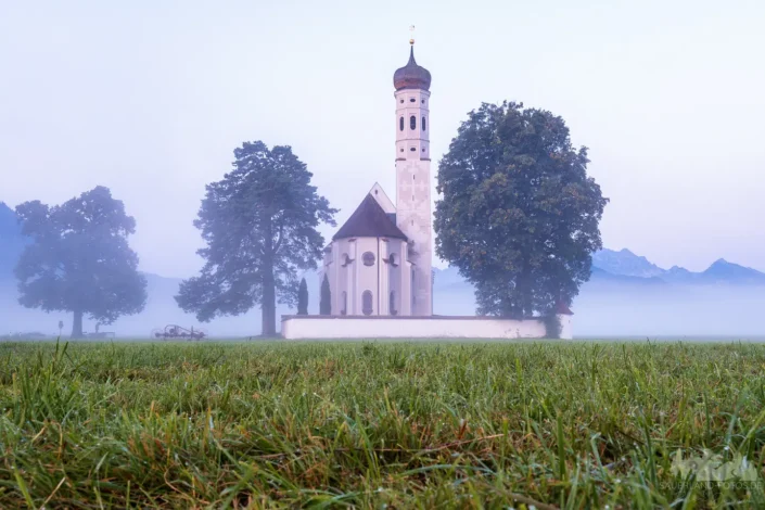 Wallfahrtskirche St. Coloman bei Schwangau im Allgäu mit taunasser Wiese im Vordergrund und dichtem Morgennebel vor den Bergen