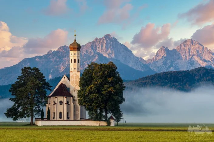 Wallfahrtskirche St. Coloman bei Schwangau mit den Gipfeln der Allgäuer Alpen, Nebel im Tal und rosa leuchtenden Wolken im Morgenlicht