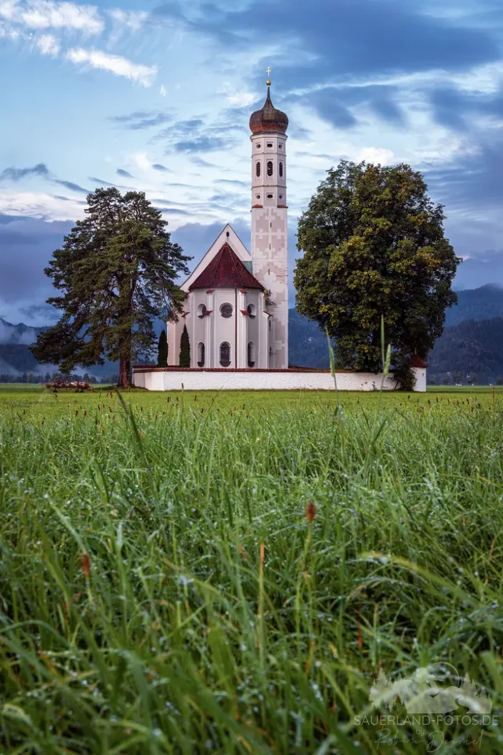 Wallfahrtskirche St. Coloman bei Schwangau im Allgäu mit taunasser Wiese im Vordergrund und Nebelschwaden vor den Bergen